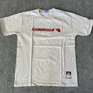 May include: A heather gray Arizona Cardinals football t-shirt. The shirt features the team name and logo in red and black across the chest. A small NFL patch is visible near the bottom hem.