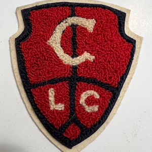 May include: A shield-shaped patch featuring red, cream, and dark blue yarn embroidery. The design includes a large cream letter C at the top, with the letters L and C in cream at the bottom, all set against a textured red background with dark blue borders.