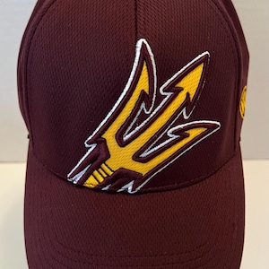 May include: A maroon baseball cap featuring a textured, breathable fabric construction. The front panel displays a prominent embroidered yellow trident logo outlined in white. The cap has a structured crown and a curved brim, with a small yellow logo visible on the side.