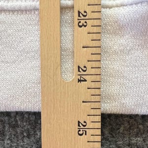 Peut inclure: Un v&ecirc;tement en tissu textur&eacute; de couleur claire est plac&eacute; derri&egrave;re une r&egrave;gle en bois. La r&egrave;gle pr&eacute;sente des marquages et des chiffres noirs nets, la section visible indiquant des mesures entre 58 et 64 centim&egrave;tres pour une r&eacute;f&eacute;rence pr&eacute;cise.