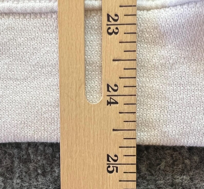 Peut inclure: Un v&ecirc;tement en tissu textur&eacute; de couleur claire est plac&eacute; derri&egrave;re une r&egrave;gle en bois. La r&egrave;gle pr&eacute;sente des marquages et des chiffres noirs nets, la section visible indiquant des mesures entre 58 et 64 centim&egrave;tres pour une r&eacute;f&eacute;rence pr&eacute;cise.