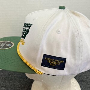 May include: White and green golf cap with the text "JOHNNIE WALKER DEVEREUX GOLF". The cap has a green brim and a yellow rope detail. A small green button is on top.