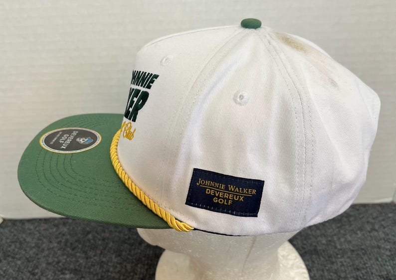 May include: White and green golf cap with the text "JOHNNIE WALKER DEVEREUX GOLF". The cap has a green brim and a yellow rope detail. A small green button is on top.