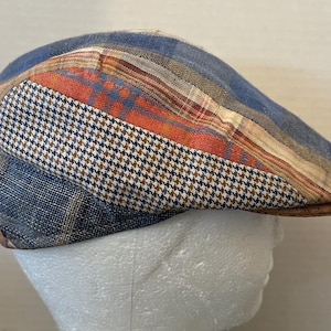 May include: A patchwork flat cap featuring a mix of patterns and colors. The cap includes blue, orange, and tan plaid and houndstooth fabrics. The brim is a solid brown color. This hat is a stylish accessory.