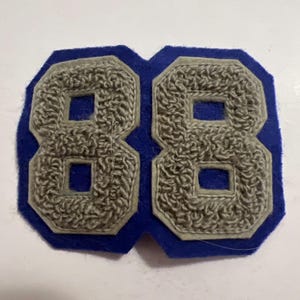 May include: A blue felt patch featuring the number 88 in a textured gray chenille material. The numbers are outlined with a light gray border and set against a solid blue felt backing that follows the contour of the double digit design.
