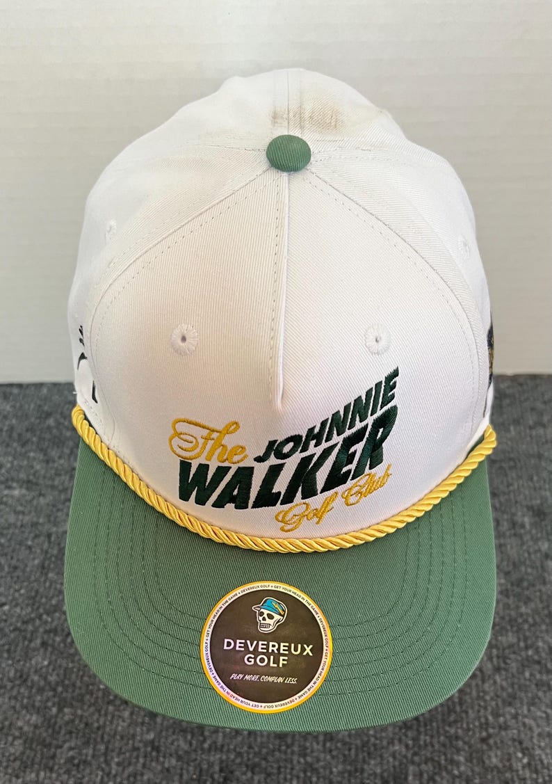 May include: White and green baseball cap with the text "The Johnnie Walker Golf Club" in gold and green. The cap has a green brim and a gold rope detail. A small green button is on top.