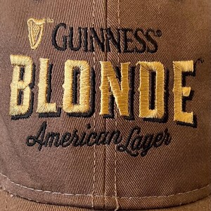 Op de afbeelding: Bruine baseballcap met het Guinness-logo en het woord "BLONDE" in gouden letters. De tekst "American Lager" is eronder geborduurd. De cap heeft een gebogen klep en een knoop aan de bovenkant.