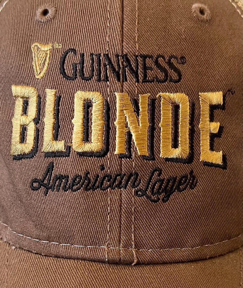 Op de afbeelding: Bruine baseballcap met het Guinness-logo en het woord "BLONDE" in gouden letters. De tekst "American Lager" is eronder geborduurd. De cap heeft een gebogen klep en een knoop aan de bovenkant.