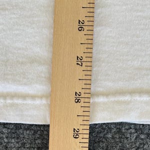 May include: A close-up of a white fabric with a wooden ruler. The ruler is marked with inch measurements from 26 to 29. The fabric appears to be a t-shirt or similar garment.