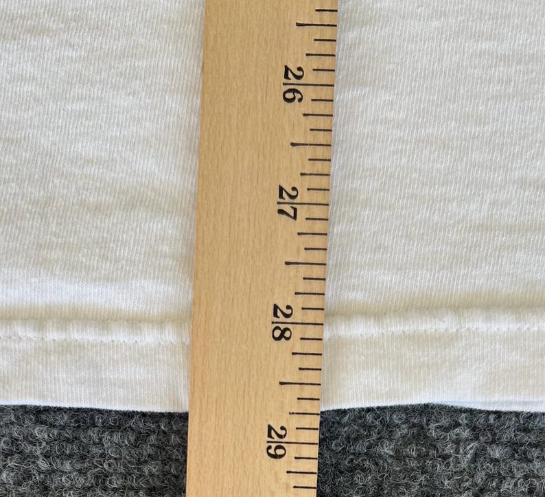 May include: A close-up of a white fabric with a wooden ruler. The ruler is marked with inch measurements from 26 to 29. The fabric appears to be a t-shirt or similar garment.
