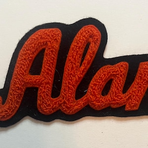 May include: A black fabric patch featuring the name Alan embroidered in a textured, bright orange thread. The lettering is in a cursive script style, and the black fabric backing is cut to follow the contour of the embroidered name.