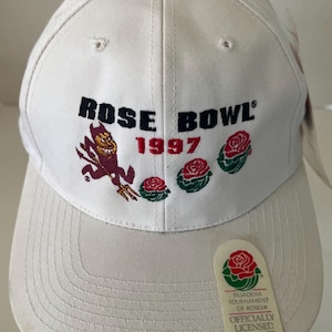 May include: A white baseball cap featuring embroidered text that reads ROSE BOWL 1997 in black and red. The design includes a maroon devil mascot and three red roses with green leaves. A tag reads PASADENA TOURNAMENT OF ROSES OFFICIALLY LICENSED PRODUCT.