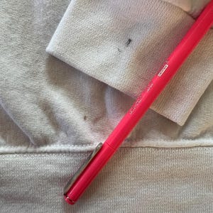 Peut inclure: Un v&ecirc;tement blanc &agrave; manches longues avec un poignet c&ocirc;tel&eacute; pr&eacute;sentant une petite tache sombre, plac&eacute; &agrave; c&ocirc;t&eacute; d'un stylo rose vif portant les inscriptions "JAPAN" et "LICHTEX" sur son corps, mettant en &eacute;vidence une tache visible sur le tissu.