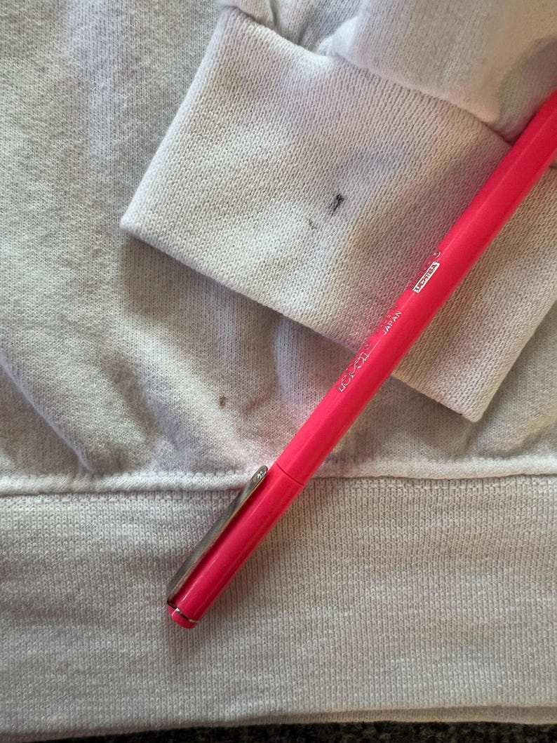 Peut inclure: Un v&ecirc;tement blanc &agrave; manches longues avec un poignet c&ocirc;tel&eacute; pr&eacute;sentant une petite tache sombre, plac&eacute; &agrave; c&ocirc;t&eacute; d'un stylo rose vif portant les inscriptions "JAPAN" et "LICHTEX" sur son corps, mettant en &eacute;vidence une tache visible sur le tissu.