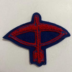 May include: A dark blue felt patch shaped like a stylized bird or arrow in flight. The patch features a raised, embroidered red outline that traces the form of a central vertical shaft with two curved wings extending outward and a tail at the base.