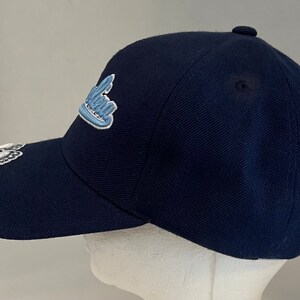 Pu&ograve; includere: Un cappello da baseball blu navy con un logo ricamato in blu chiaro e bianco che dice "California".