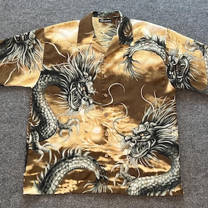 May include: A short-sleeve button-down shirt in shades of brown and gold featuring a repeating dragon print. The shirt has a classic collar, a front button placket with five visible buttons, and a label at the neck reading No Boundaries.