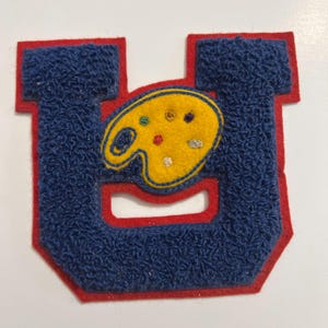 May include: A varsity-style letter U patch featuring a textured blue chenille surface with a red felt border. Centered on the letter is a yellow felt artist palette shape with small embroidered paint spots in green, brown, red, and white colors.