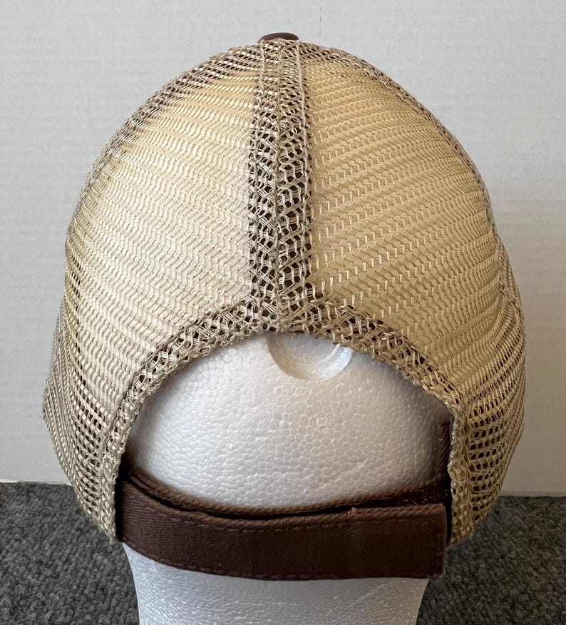 Op de afbeelding: Een bruine en beige truckerpet, van achteren gezien, met een mesh rug en een effen voorkant. De pet heeft een bruine verstelbare band en een bruine knoop aan de bovenkant. De mesh is beige en de effen voorkant is bruin.