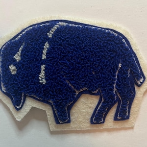 May include: A blue punch needle patch in the shape of a buffalo, featuring a textured loop pile surface and white stitched outlines. The buffalo figure is set against a cream-colored felt backing that follows the contour of the animal shape.