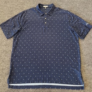 May include: Navy blue polo shirt with a white geometric pattern. The shirt has short sleeves, a collared neckline, and a three-button placket. A white stripe accents the bottom hem.