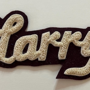 May include: A dark maroon felt patch featuring the name Larry embroidered in a cream-colored, textured yarn. The lettering is in a cursive script style, with the cream yarn creating a raised, looped appearance against the dark background.