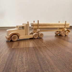 Könnte beinhalten: Ein hölzerner Spielzeug-LKW mit einem Anhänger, der Holzstämme transportiert. Der LKW und der Anhänger sind aus Naturholz gefertigt und haben ein rustikales Aussehen. Der LKW hat vier Räder und der Anhänger sechs Räder.