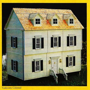 May include: A white dollhouse with a brown shingled roof and black shutters. The dollhouse has three floors and a front porch with white railings. The text "Yorktown Colonial" is printed below the dollhouse.