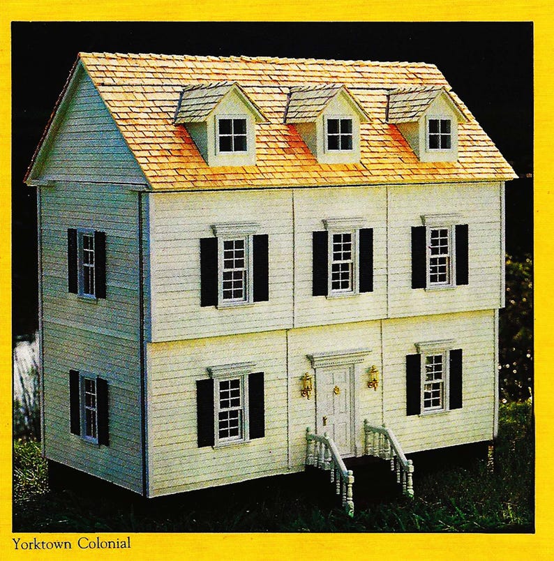 May include: A white dollhouse with a brown shingled roof and black shutters. The dollhouse has three floors and a front porch with white railings. The text "Yorktown Colonial" is printed below the dollhouse.