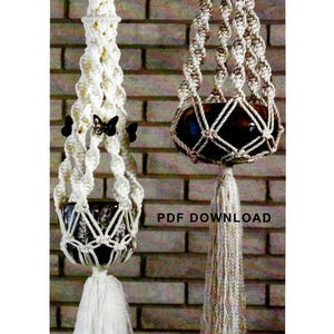 May include: Two macrame plant hangers with a brown pot in each. The plant hangers are made with white and beige cord and have a tassel at the bottom. The hangers are hanging from a brick wall. The text "PDF DOWNLOAD" is visible in the image.