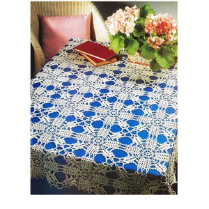 May include: A white crocheted tablecloth with a floral pattern covers a blue table. A red book rests on the table, near a wicker chair with a pink pillow. A basket of pink and green flowers sits on the table.