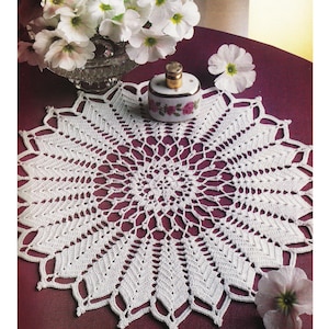 May include: White crocheted doily with a delicate floral pattern. The doily is laid on a burgundy surface with white flowers and a small porcelain bottle with a floral design.