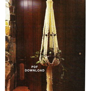 May include: A white macrame plant hanger with a tassel hanging from the bottom. The hanger is hanging from a ceiling in a room with a wooden door and a stone wall. The text "PDF DOWNLOAD" is visible in the image.