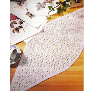 Könnte beinhalten: Weißer, offen gewebter, gehäkelter Tischläufer mit sich wiederholendem geometrischem Muster. Der Läufer liegt auf einer Holzoberfläche, mit Schreibwaren mit floralen Motiven und einer Teetasse in der Nähe. Das Design des Läufers ist symmetrisch und filigran.