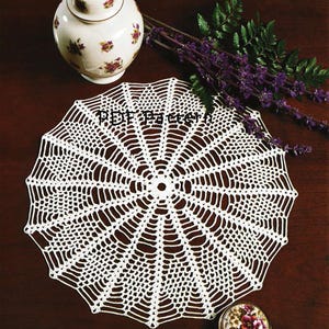 May include: A white, crocheted doily with a spiderweb design, a small floral jar, and a decorative jar with a lid. The text "PDF Pattern" is visible on the doily. Purple flowers and greenery are also present.