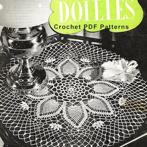 Op de afbeelding: Een vintage boek met patronen voor gehaakte doilies met een witte gehaakte doily op een donker oppervlak. Het boek is getiteld "Doilies Crochet PDF Patterns" en bevat de tekst "A Collection of Fascinating New Designs".