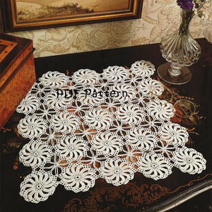 Puede incluir: Doily de ganchillo blanco con un patrón floral. El doily está sobre una mesa de madera oscura con un jarrón de flores moradas y una imagen enmarcada en el fondo.