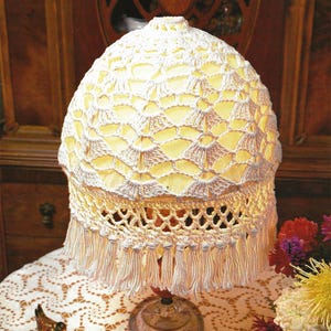May include: A white crocheted lampshade with a fringe trim sits on a silver base on a white crocheted tablecloth. The lampshade is lit and casts a warm glow.
