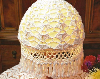 Puede incluir: Una pantalla de lámpara de crochet blanco con un borde de flecos se asienta sobre una base plateada sobre un mantel de crochet blanco. La pantalla de la lámpara está encendida y emite un brillo cálido.