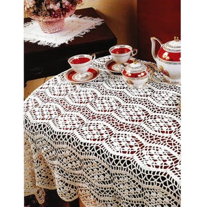 May include: A white, crocheted lace tablecloth covers a round table, with a red surface visible through the openwork design. Two teacups and saucers, a sugar bowl, and a teapot with red and gold accents are arranged on the table. A basket of flowers sits in the background.