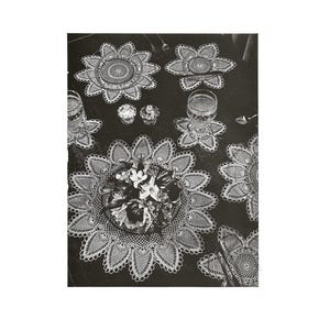 May include: A black and white photograph of a table setting. The table is set with several white, crocheted doilies in a floral pattern. There are also glasses, spoons, and a centerpiece of flowers.