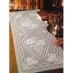 May include: A white, intricately crocheted table runner with floral patterns. The rectangular runner features detailed openwork designs and a scalloped edge. The runner is displayed on a dark wooden surface, showcasing its delicate craftsmanship.