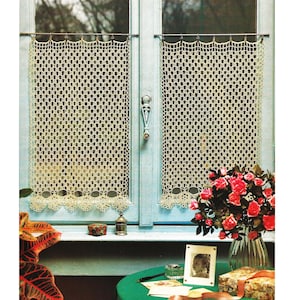 May include: A window with light blue trim features two crocheted lace curtains. A vase of red roses sits on a table covered with a green cloth. Other items on the table include a framed photo, a small box, and a decorative container.