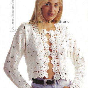 Puede incluir: Chaqueta de ganchillo blanca con un patrón de encaje y un cierre de botón. La chaqueta tiene un borde festoneado y se muestra puesta con jeans azules.