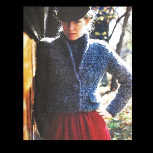 May include: A woman wearing a dark gray textured jacket with a single button closure and a small pocket. The jacket is paired with a red skirt and a black hat. The background features a wooden wall and trees.