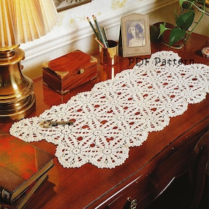 May include: A cream-coloured, openwork crochet table runner with a floral pattern, displayed on a polished wooden desk. The runner has a scalloped edge. A vintage key and books are also visible.