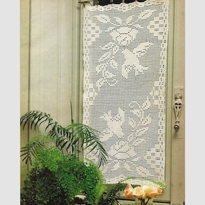 May include: A white, crocheted curtain with a floral and bird design hangs in a doorway. The curtain is rectangular and has a detailed, openwork pattern. Green plants and flowers are in front of the curtain.
