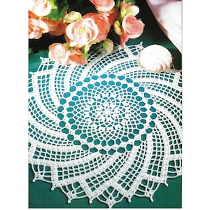 Peut inclure: Napperon blanc en crochet avec un motif en spirale. Le motif de dentelle complexe présente un motif floral central et un bord festonné. Le napperon est présenté sur une surface turquoise, avec des fleurs couleur pêche en arrière-plan.