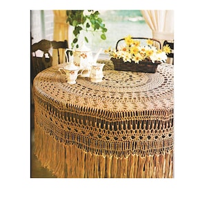 May include: A circular, crocheted tablecloth in a light brown hue, featuring a fringed border. It drapes over a dark wooden table. A teapot, creamer, sugar bowl, and two teacups are arranged on the table. A basket of yellow flowers is also present.
