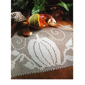 Könnte beinhalten: Weißer, gehäkelter Tischläufer mit Kürbismuster. Der Läufer liegt auf einer Holzoberfläche, mit einer Schüssel Gemüse, einem Krug und einem karierten Tuch im Hintergrund. Das Design des Läufers zeigt eine große Kürbisform.